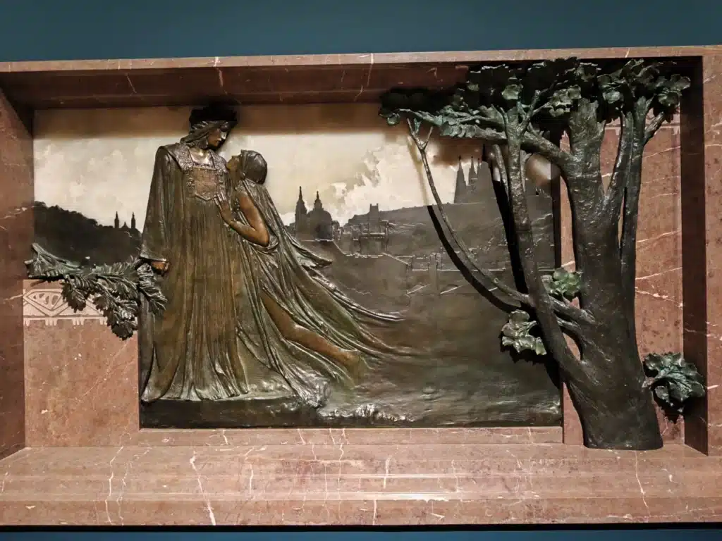 Bronze relief by Stanislav Sucharda depicting figures symbolizing Prague and the Vltava River, artwork from Flowing Through Czech Culture – The Vltava Exhibition in Prague, featured on SOLARIS Language School blog about Czech culture and learning Czech.
